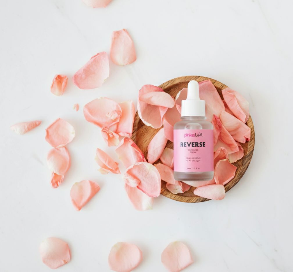 Skincare bottle labeled 'REVERSE' on a wooden plate with pink flower petals on a light background