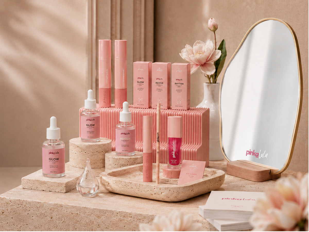 A styled beauty product display featuring pink cosmetic bottles, tubes, and boxes arranged on stone pedestals beside a rounded mirror with a gold frame. The products include pink liquid and cream items with droppers and applicators, set against a warm beige background with soft floral decor and leaf shadows.