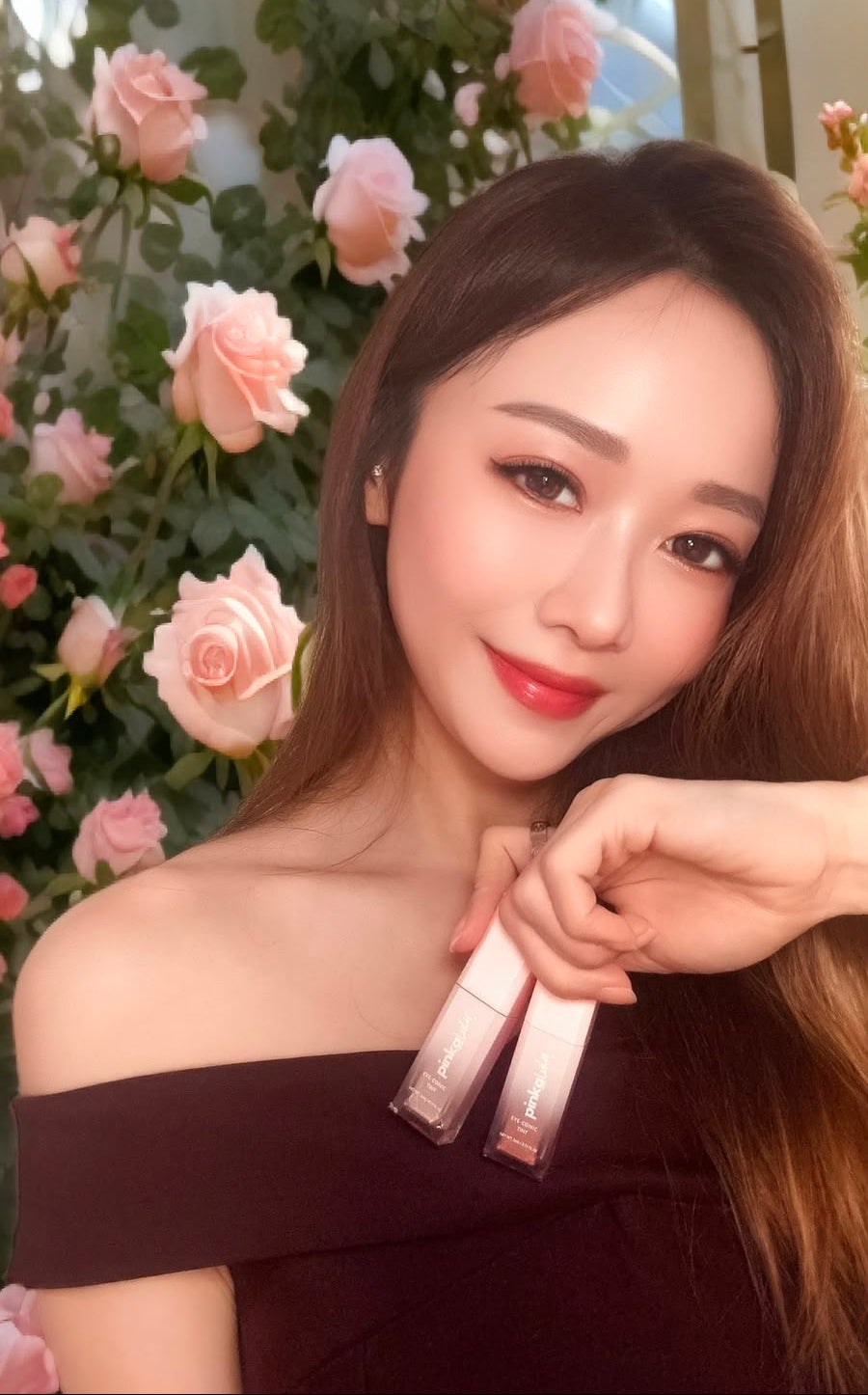 A woman with long light brown hair poses in front of a floral wall of pink roses, holding two tubes of Pinkalulu Eye-conic Tint.